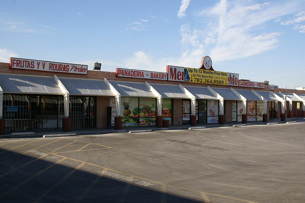 A single-storey Las Vegas shopping plaza in hard afternoon light: a row of shopfronts under white metal awnings with hand-painted signs reading Frutas y Verduras, Panadería, and Carnicería, and an empty parking lot in front.